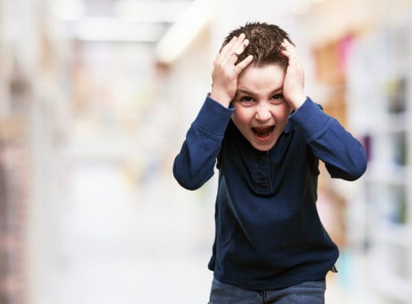 Young boy in a blue shirt holding his head with both hands and shouting in frustration indoors