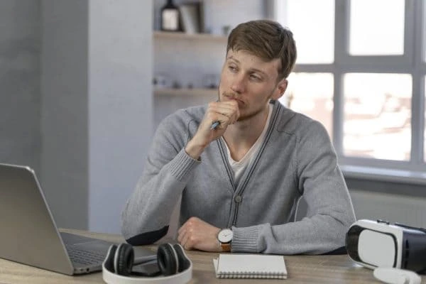 Man sitting at a desk with a laptop and headphones looking thoughtful